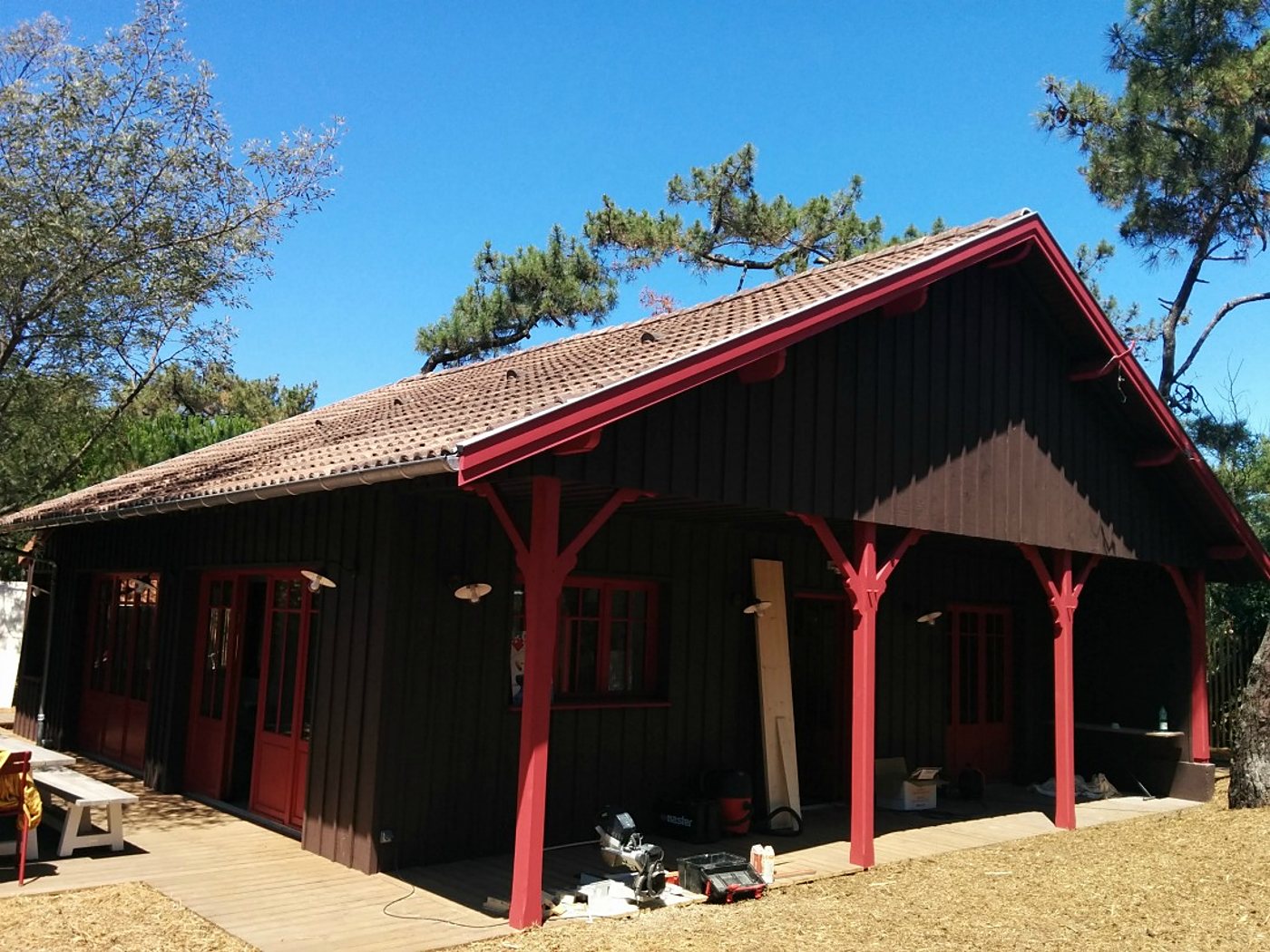Une construction en bois brun foncé aux poteaux et menuiseries rouges, sous des pins, avec une scie à onglet et des outils posés sur la terrasse.