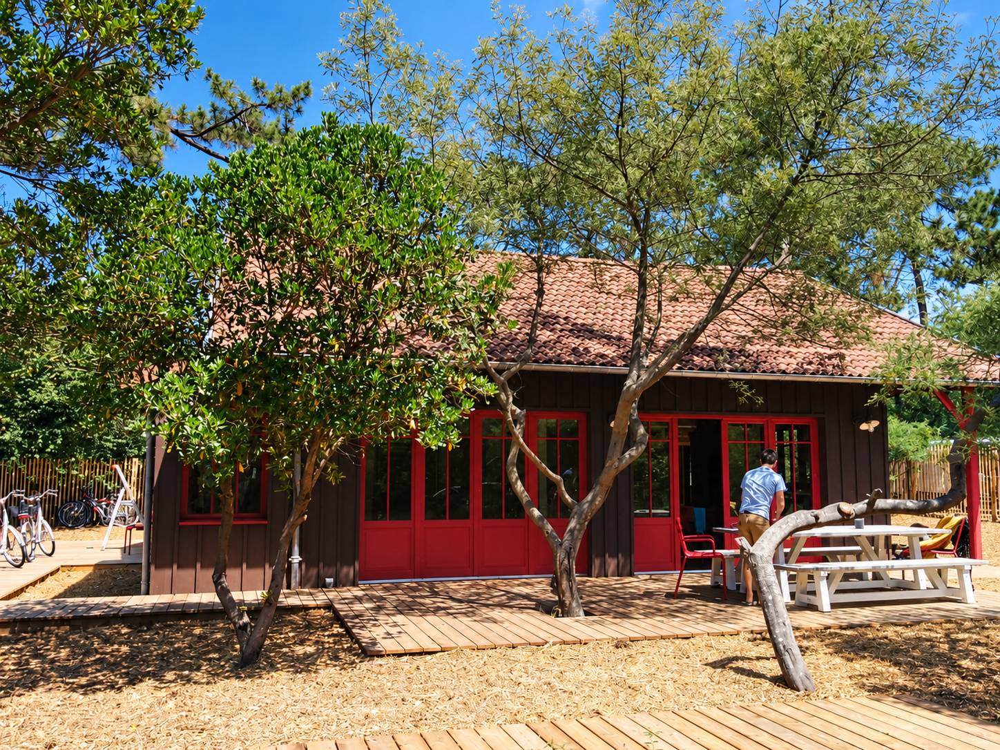 Une cabane en bois sombre aux grandes portes rouges, entourée d'arbres, avec une terrasse en bois, une table blanche et des vélos garés à gauche.