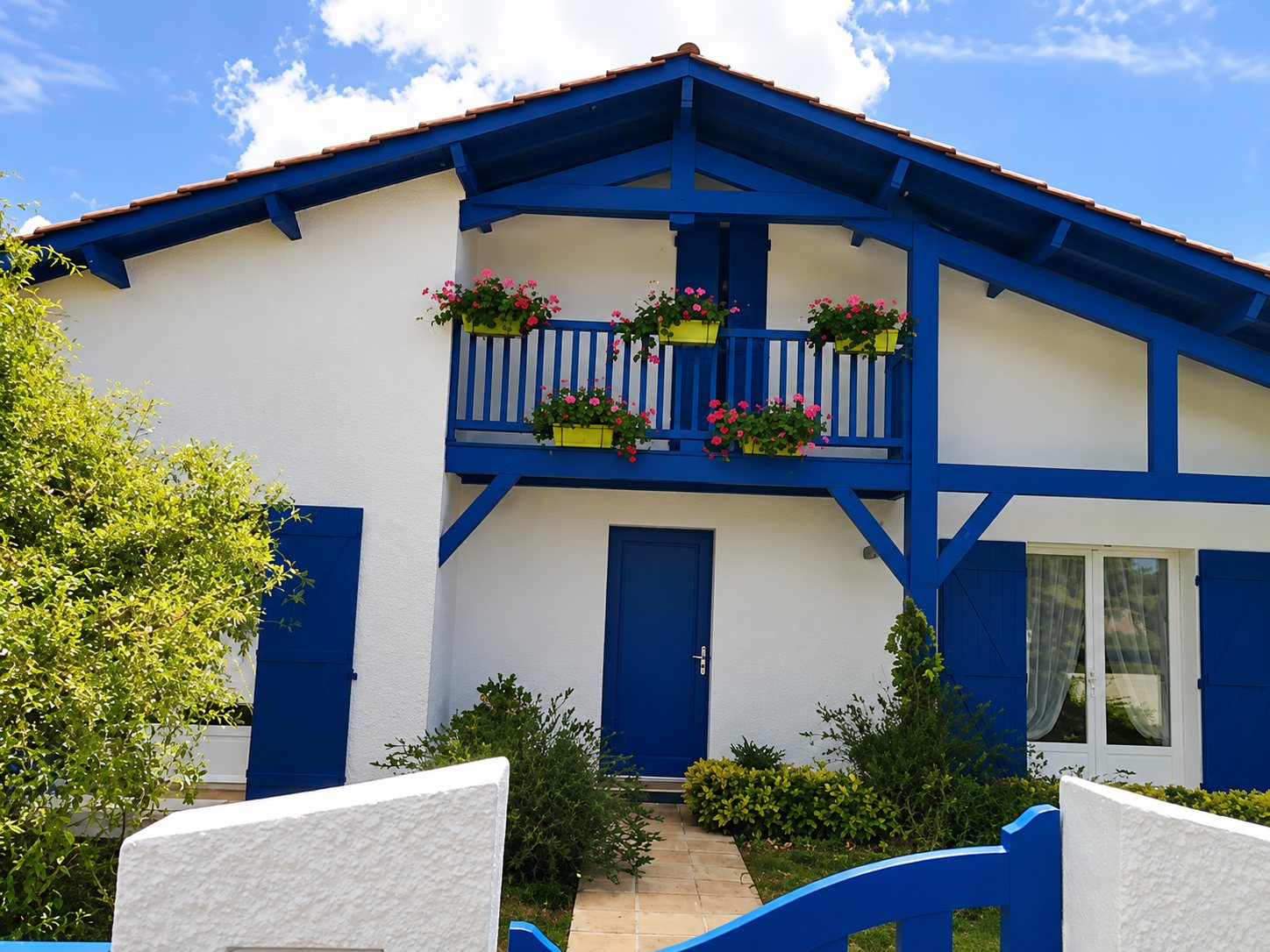 Façade d'une maison blanche aux boiseries et volets bleus, avec un balcon orné de jardinières de fleurs et un petit jardin devant.