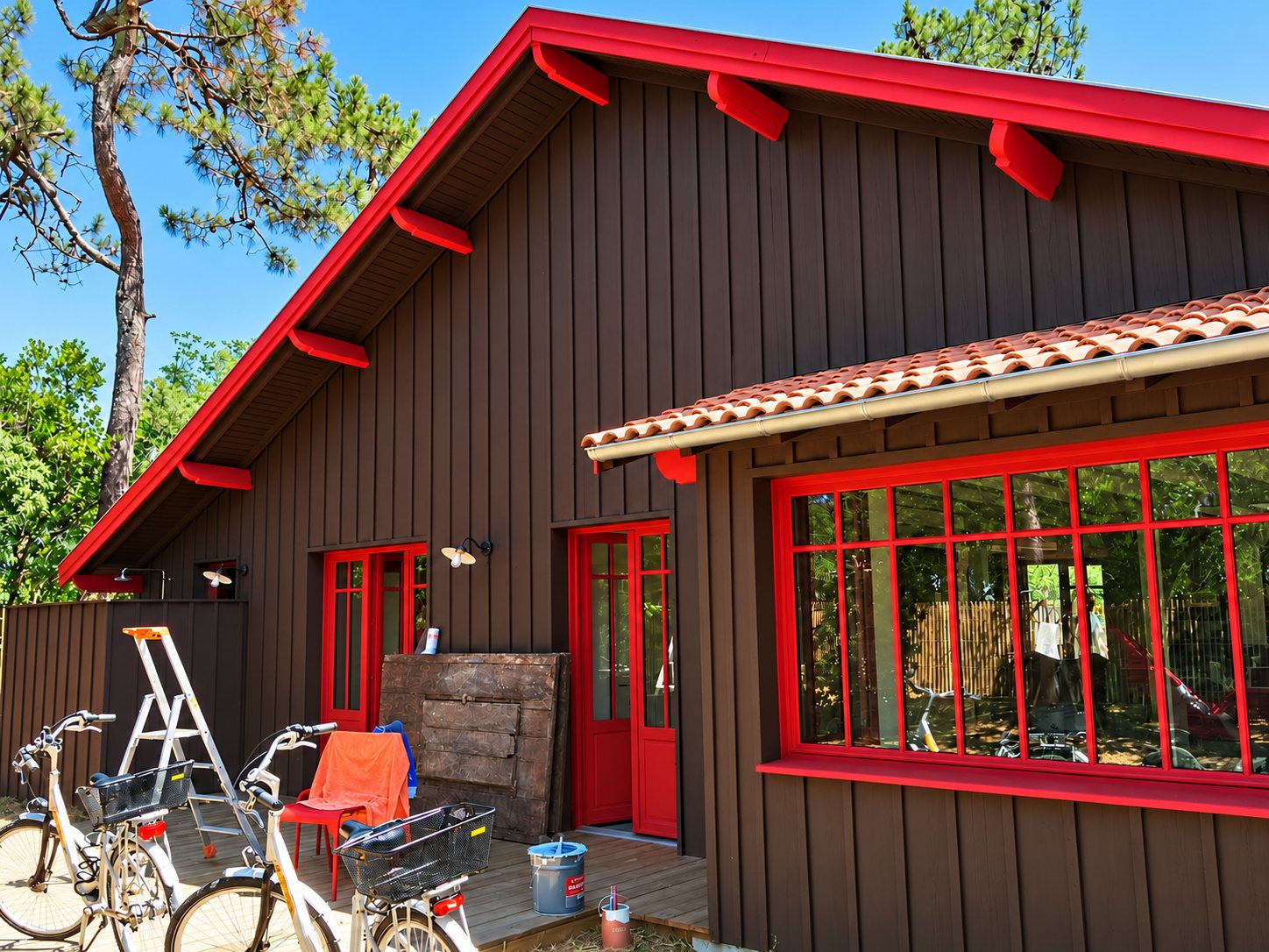 Façade d'une maison en bois brun aux boiseries et fenêtres rouges, avec toit de tuiles, vélos et escabeau sur la terrasse, entourée de pins.