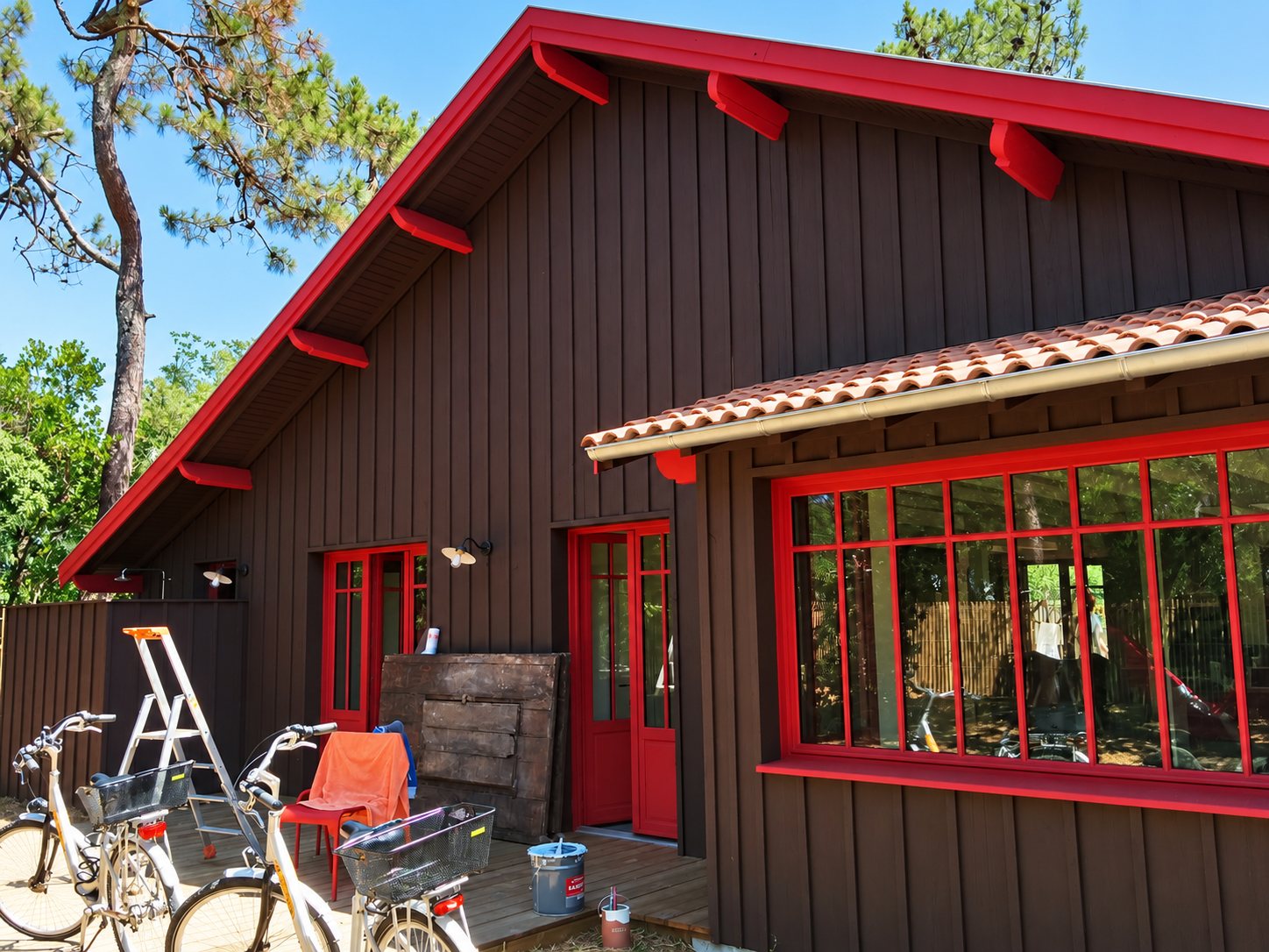 Façade d'une maison en bois brun aux boiseries et fenêtres rouges, avec toit de tuiles, vélos et escabeau sur la terrasse, entourée de pins.