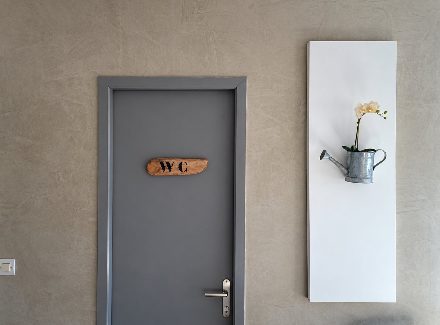 Une porte grise portant un panneau en bois indiquant WC, à côté d'un panneau blanc sur lequel est accroché un petit arrosoir en zinc contenant une orchidée.