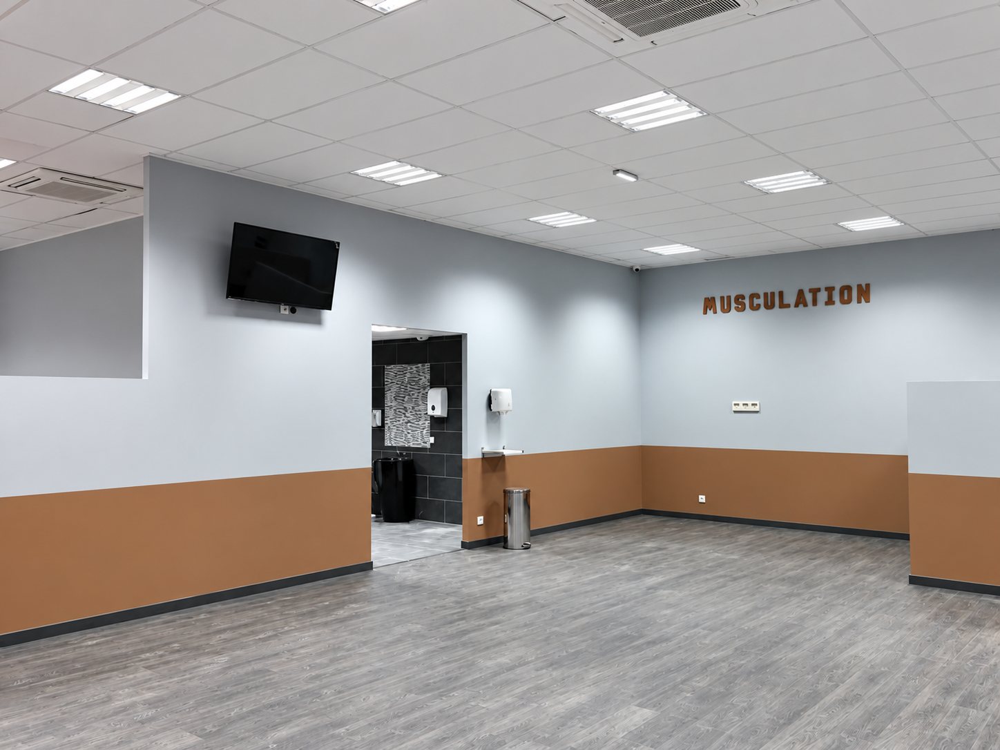 Grande salle vide aux murs gris clair et marron, avec un écran fixé au mur, un plafond lumineux et le mot MUSCULATION affiché au fond.